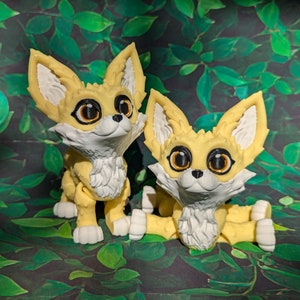 Two cute 3D printed fennec foxes sit in the grass backdrop. They are yellow and white with black and gold eyes. They have huge ears, big eyes, and a clever look on their faces.