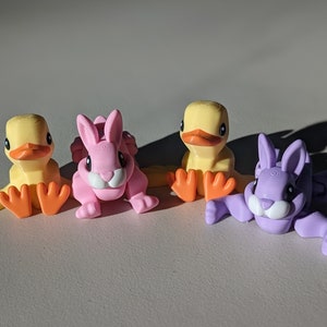 May include: Four small plastic figurines of animals. A yellow duck, a pink rabbit, and two purple rabbits. The rabbits are in a sitting position, while the duck is standing with its wings out.