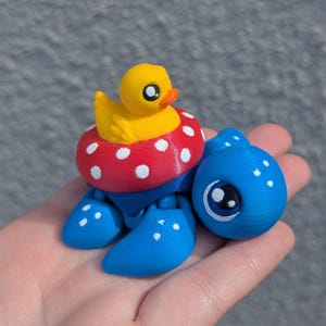 May include: A whimsical 3D-printed toy featuring a yellow duck in a red and white polka-dot inner tube, atop a blue creature with large eyes. The toy is held in a hand, showcasing its playful design.