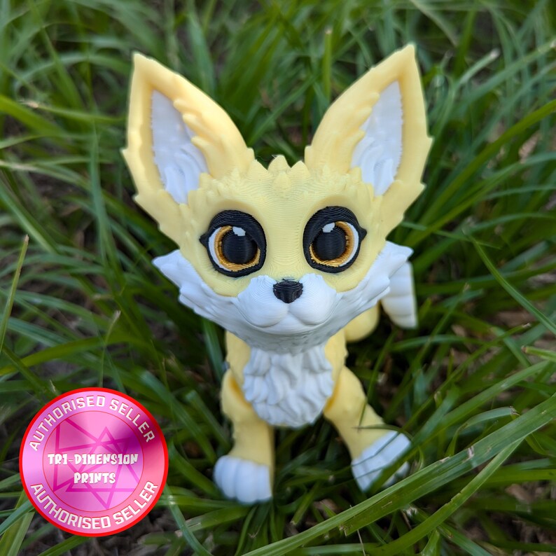 A cute 3D printed fennec fox sits in the grass. It is yellow and white with black and gold eyes. It has huge ears, big eyes, and a clever look on its face.