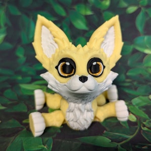 A cute 3D printed fennec fox with its legs splayed out against a forest backdrop. It is yellow and white with black and gold eyes. It has huge ears, big eyes, and a clever look on its face.
