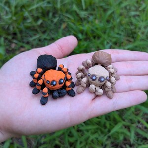 May include: Two small, handmade spider figurines. One is orange and black, the other is tan and brown. The spiders have detailed legs and eyes, and are likely made of clay or a similar material. The figurines are displayed on a hand.