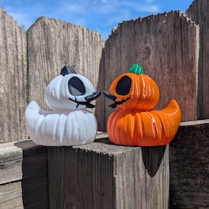 May include: Two Halloween-themed duck figurines. One is white with a ghost-like appearance, and the other is orange, resembling a pumpkin. Both have black eyes and beaks, and are perched on a wooden surface against a blue sky.
