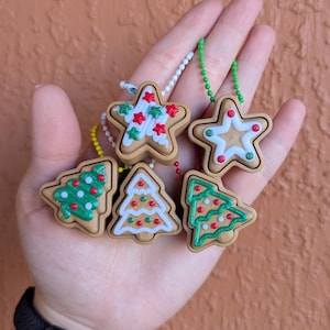 May include: A collection of Christmas-themed ornaments in the shape of gingerbread cookies. The ornaments include stars and Christmas trees, decorated with white icing and red and green accents. Each ornament is attached to a beaded chain.