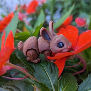 May include: A small, brown, articulated rabbit figurine with black eyes is nestled in a red flower. The rabbit is made of a textured material and has a realistic appearance.