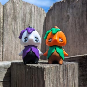May include: Two small, whimsical figurines. One is white with a purple hat and vest, black pants, and small arms. The other is orange with a green leafy collar and hat, brown body, and small arms. Both have large, expressive eyes.