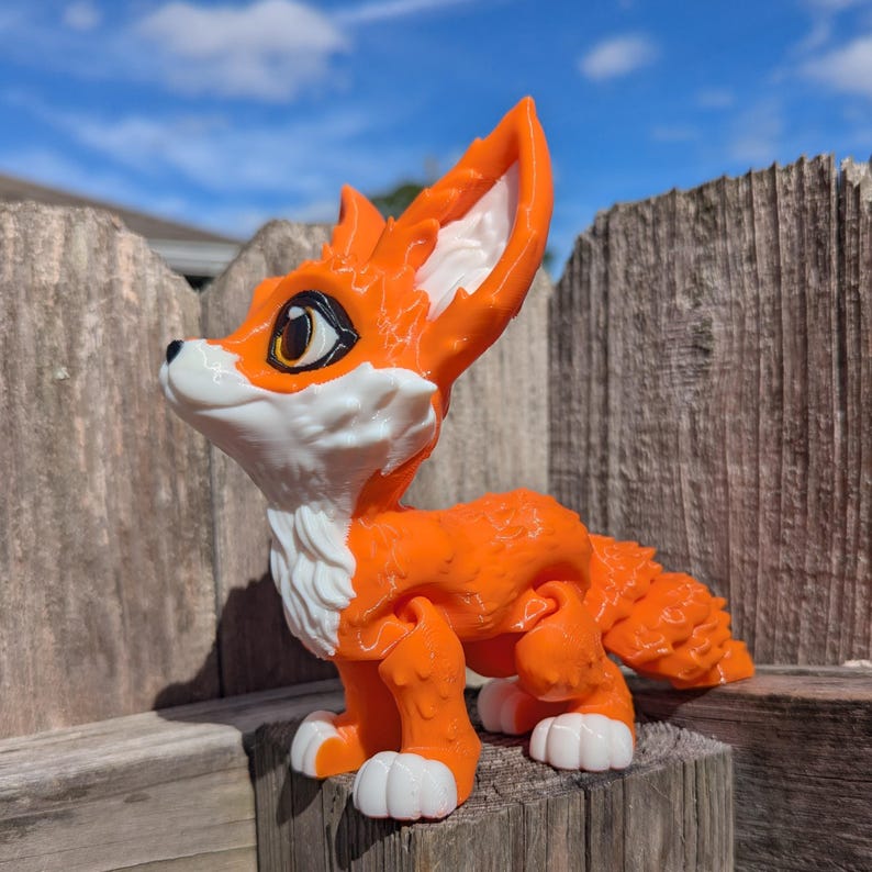 A cute 3D printed fennec fox sits on a fence post. It is orange and white with black and gold eyes. It has huge ears, big eyes, and a clever look on its face.