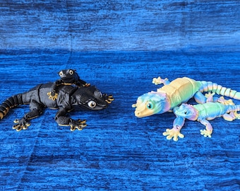May include: Two toy lizards are displayed on a blue surface. One is black with gold accents and a smaller lizard on its back. The other two lizards are pastel rainbow colors. The lizards are a fun toy.