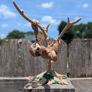 May include: A brown sloth figurine hanging from a tree branch. The sloth has large, round eyes and is suspended from a light brown tree with green accents at the base. The background includes a wooden fence and a blue sky.