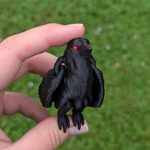 May include: A black figurine of a bird-like creature with red eyes and outstretched wings. The figure has a textured surface and is held in a hand against a blurred green background.