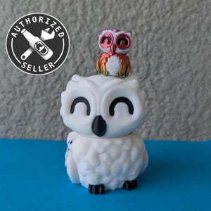 May include: A white ceramic owl figurine with black eyes and a small pink and red owl figurine perched on its head.