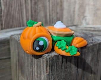 May include: A collection of 3D-printed fall-themed decorations. Includes an orange pumpkin with a cartoon eye, a slice of pie with whipped cream, and a small carrot with green leaves. The items are arranged on a wooden surface.