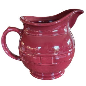 May include: A deep burgundy ceramic pitcher featuring a rounded body with a raised basket-weave texture around the center, a curved handle on the left side, and a wide pouring spout at the top rim, finished with a smooth, glossy glaze.