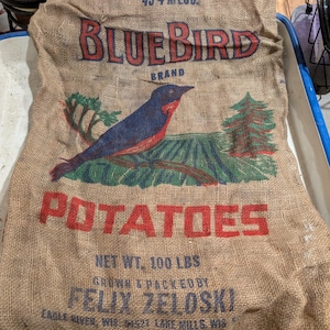 May include: A burlap potato sack with the words "BLUEBIRD BRAND POTATOES" in red and blue lettering. The sack features an illustration of a bluebird and a field. The net weight is 100 lbs.