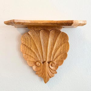 May include: A wooden wall shelf with a carved seashell design. The shelf is made of light brown wood and features a rectangular top with a decorative, fan-shaped support. The seashell design has detailed lines and curves.