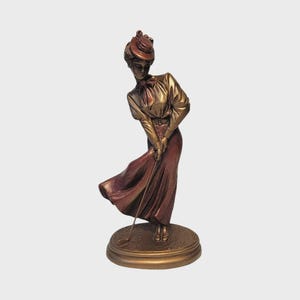 May include: Bronze-colored sculpture of a woman in a long skirt and hat, poised to swing a golf club. The figure is on a circular base. The woman is wearing a long skirt and a blouse with a bow.