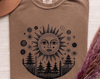 May include: A brown t-shirt featuring a black graphic of a sun with a face, surrounded by stars and trees. The graphic is enclosed in a circle.