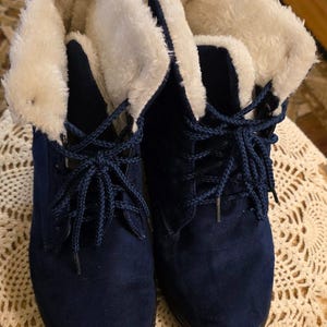 May include: A pair of navy blue suede ankle boots with cream-colored faux fur lining. The boots have dark blue laces and black soles. The boots are designed for cold weather and are suitable for casual wear.