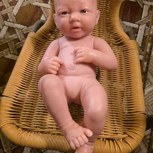 May include: A lifelike baby doll with a realistic skin tone, sitting in a woven wicker chair. The doll has detailed facial features, including blue eyes and a slightly open mouth. The doll is nude and has articulated limbs.