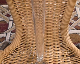 May include: A tall, clear glass vase with a round, textured base and a long, cylindrical neck. The vase is transparent, allowing light to pass through. It sits against a woven wicker chair.