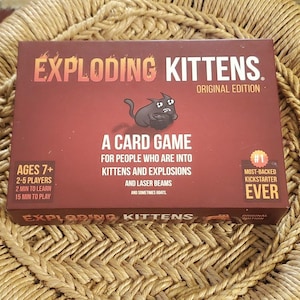 May include: A boxed card game titled "Exploding Kittens" in a red and brown color scheme. The box features a cartoon cat and text that reads "A Card Game for people who are into kittens and explosions and laser beams." The game is for ages 7+.