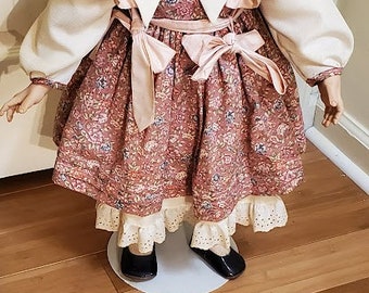 May include: A porcelain doll wearing a pink and white dress with a white collar and pink bow. The doll has blonde hair and blue eyes.
