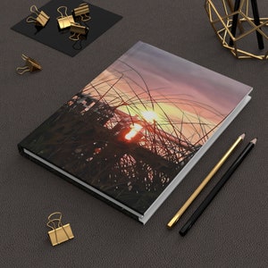 May include: A white hardback journal with a colourful sunset photo on the cover. The photo features tall grass silhouetted against a pink and orange sky with a bright sun shining through the grass.