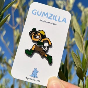 May include: A green, yellow, and brown enamel pin of a cartoon character running with a green plant in their hand. The pin is on a white card with the text "GUMZILLA -hard enamel pin- @gumzillashop".
