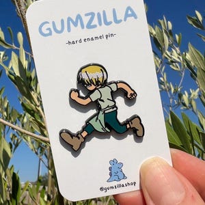 Può includere: Un cartoncino bianco con il testo "GUMZILLA -hard enamel pin- " e un'immagine di un cartone animato di una persona con i capelli biondi che corre. L'immagine è una spilla in smalto duro. Sotto l'immagine c'è un piccolo dinosauro di cartone animato blu con il testo "@gumzillashop".