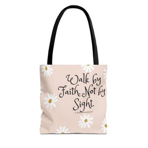 May include: A tote bag with a light pink background and a daisy pattern. The bag features the text "Walk by Faith, Not by Sight" in black script. The bag has black handles.