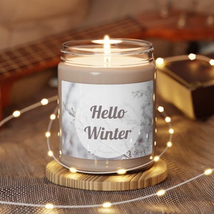 Pode incluir: Uma vela marrom com um rótulo branco que diz "Hello Winter". A vela está sentada em um descanso de vela de madeira e está cercada por luzes de corda brancas.