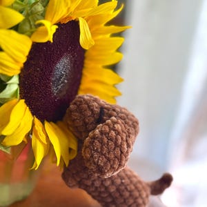May include: A small brown crocheted plush dog with a textured yarn surface, positioned next to a large, bright yellow sunflower with a dark brown center. The dog has floppy ears and a small tail, crafted from soft, plush material.