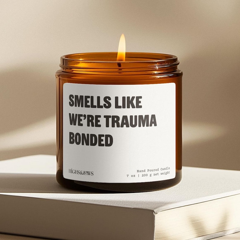 May include: A lit, brown glass candle jar with a white label that reads "SMELLS LIKE WE'RE TRAUMA BONDED." The candle is labeled "HIGHS&JOWS" and states it is a hand-poured candle, 7 oz / 200 g net weight.