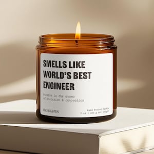 May include: A brown glass candle jar with a white label that reads "Smells Like World's Best Engineer". The label also includes the text "Breathe in the aroma of precision & innovation" and "HIGHS&LOWS". The candle is lit and the jar is sitting on a white surface.