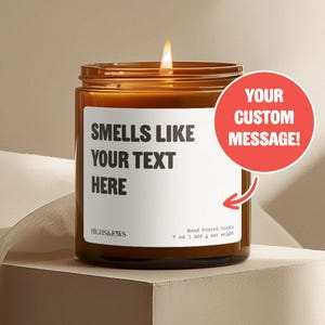 Peut inclure: Une bougie en verre brun allumée avec une étiquette blanche indiquant "SMELLS LIKE YOUR TEXT HERE". Un cercle rouge avec le texte "YOUR CUSTOM MESSAGE!" est à droite. La bougie pèse 200 g net.