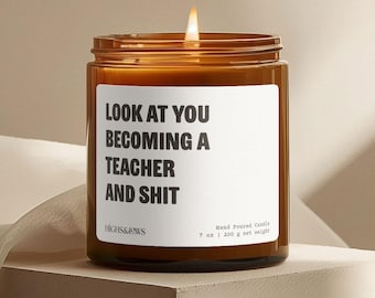 May include: A lit, hand-poured candle in a brown glass jar. The white label reads "LOOK AT YOU BECOMING A TEACHER AND SHIT." The candle weighs 7 oz (200 g). The brand name "HIGHS&LOWS" is also visible.