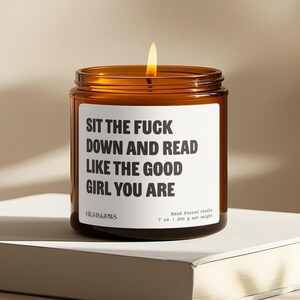 May include: A lit, amber-colored glass jar candle with a white label. The label reads "SIT THE FUCK DOWN AND READ LIKE THE GOOD GIRL YOU ARE." The candle is hand-poured and weighs 7 oz (200 g).