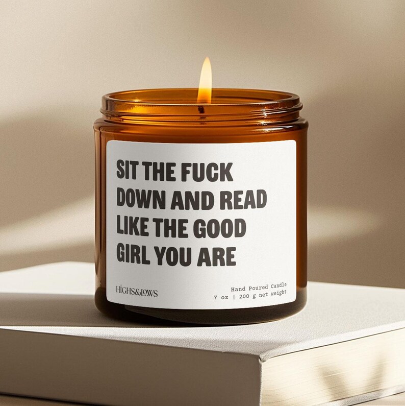 May include: A lit, amber-colored glass jar candle with a white label. The label reads "SIT THE FUCK DOWN AND READ LIKE THE GOOD GIRL YOU ARE." The candle is hand-poured and weighs 7 oz (200 g).