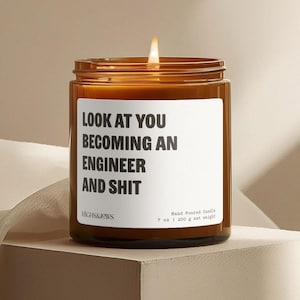 Könnte beinhalten: Ein brennendes, braunes Glas-Kerzenglas mit einem weißen Etikett mit dem Text "LOOK AT YOU BECOMING AN ENGINEER AND SHIT". Die Kerze ist handgegossen und wiegt 200 g (7 oz).
