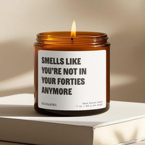 May include: A brown glass candle with a white label that reads "Smells Like You're Not In Your Forties Anymore". The candle is 7 ounces and weighs 200 grams.
