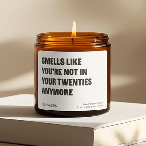 May include: A lit, amber-colored glass candle jar with a white label. The label reads "SMELLS LIKE YOU'RE NOT IN YOUR TWENTIES ANYMORE." The candle is hand-poured and weighs 7 oz (200 g).