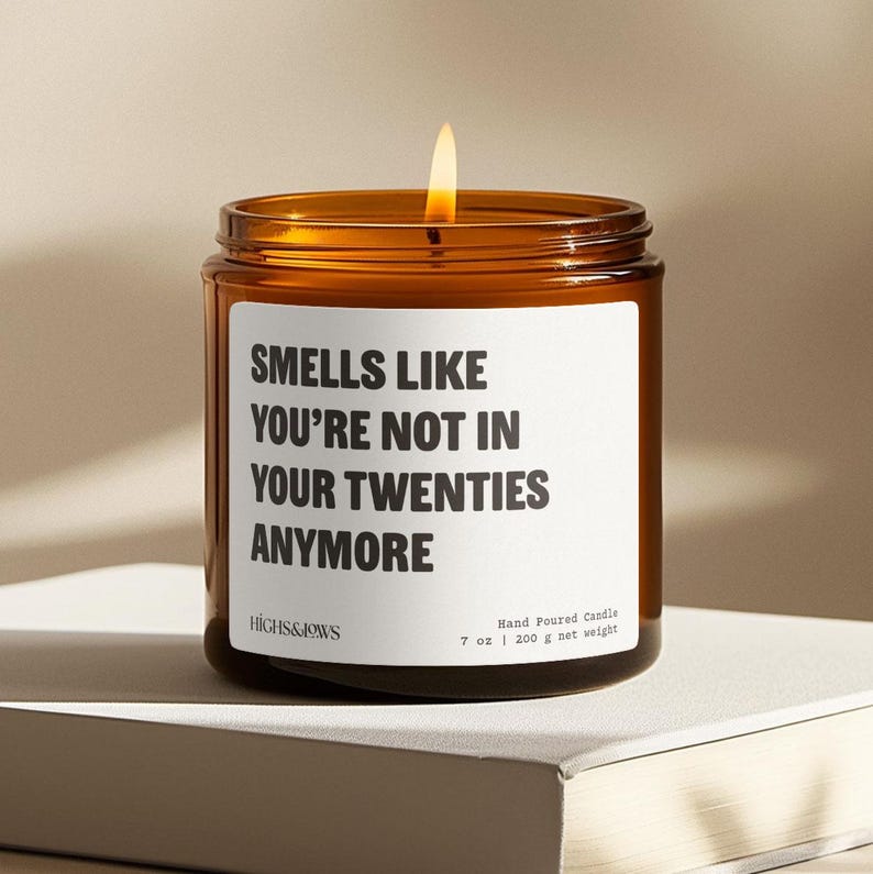 May include: A lit, amber-colored glass candle jar with a white label. The label reads "SMELLS LIKE YOU'RE NOT IN YOUR TWENTIES ANYMORE." The candle is hand-poured and weighs 7 oz (200 g).