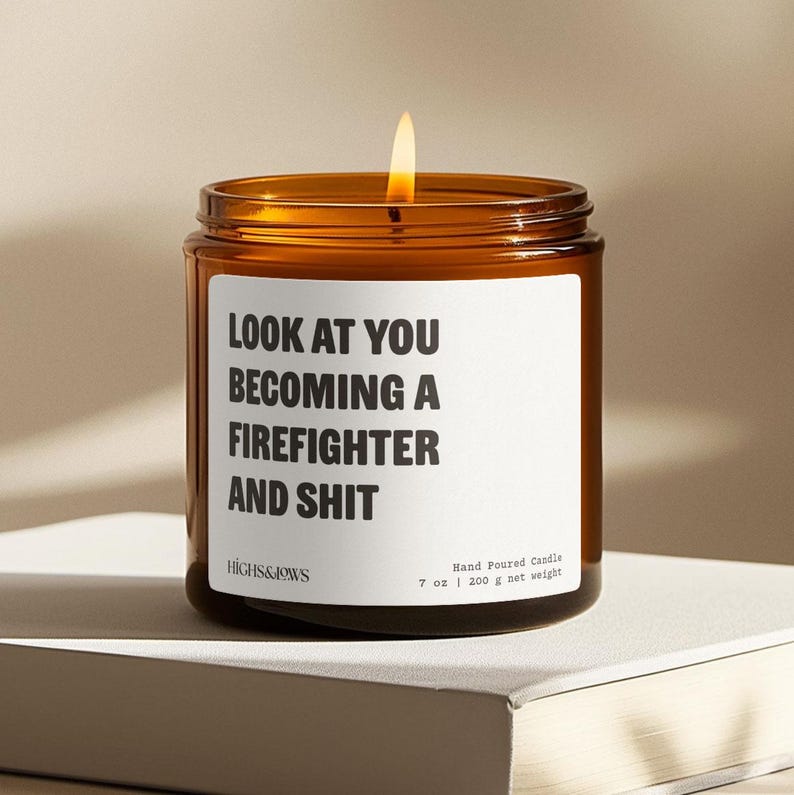 以下が含まれることがあります： 白いラベルに「LOOK AT YOU BECOMING A FIREFIGHTER AND SHIT」と書かれた茶色のガラス製のキャンドル。キャンドルに火が灯っており、ラベルにはブランド名「HIGHS&JOWS」と「Hand Poured Candle 7 oz | 200 g net weight」の文字も記載されています。
