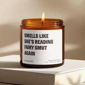 Peut inclure: Une bougie en verre marron allumée avec une étiquette blanche. L'étiquette indique "SMELLS LIKE SHE'S READING FAIRY SMUT AGAIN". La bougie est coulée à la main et pèse 200 g.