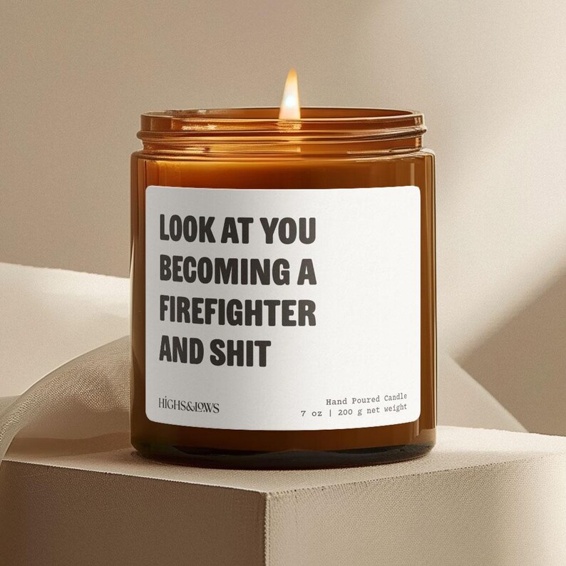 以下が含まれることがあります： 白いラベルに「LOOK AT YOU BECOMING A FIREFIGHTER AND SHIT」と書かれた茶色のガラス製のキャンドル。キャンドルに火が灯され、白い芯が見えます。ラベルにはブランド名「HIGHS&JOWS」と「Hand Poured Candle 7 oz | 200 g net weight」の文字も書かれています。