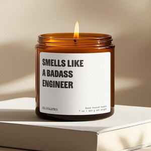 May include: A brown glass candle jar with a white label that reads "Smells Like A Badass Engineer". The candle is lit and the jar is sitting on a white surface.