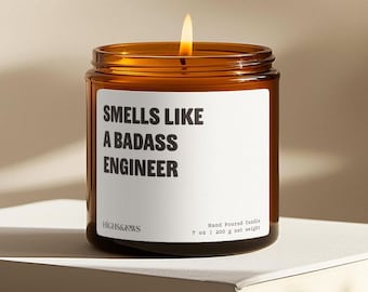May include: A brown glass candle jar with a white label that reads "Smells Like A Badass Engineer". The candle is lit and the jar is sitting on a white surface.