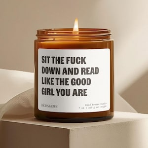 May include: A lit, hand-poured candle in a brown glass jar. The white label reads "SIT THE FUCK DOWN AND READ LIKE THE GOOD GIRL YOU ARE." The candle is 7 oz (200 g) net weight.