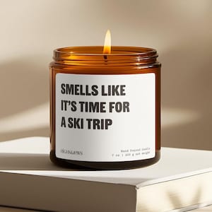 May include: A brown glass candle with a white label that reads "Smells Like It's Time For A Ski Trip". The candle is lit and the label also includes the brand name "HIGHS&JOWS" and the text "Hand Poured Candle 7 oz | 200 g net weight".