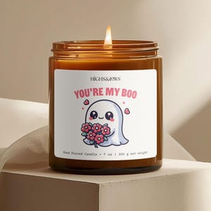 Może przedstawiać: Brązowa świeca w szklanym słoiku z białą etykietą, na której widnieje napis "HIGHS&LOWS YOU'RE MY BOO". Etykieta przedstawia kreskówkowego ducha trzymającego różowe kwiaty. Świeca jest ręcznie wylewana i waży 7 uncji lub 200 gramów.